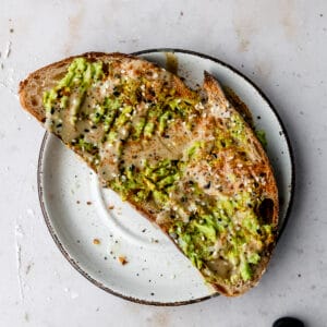 slice of tahini avocado toast on a white plate topped with old bay and everything bagel seasoning