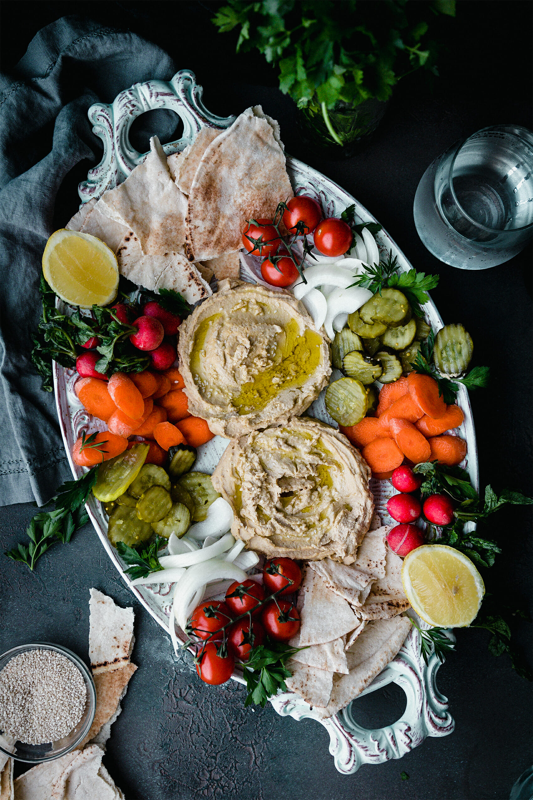 platter of creamy ottolenghi hummus surrounded by carrots, pickles, onions, pita, and cherry tomatoes with fresh herbs