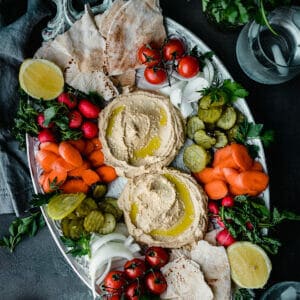 platter of creamy ottolenghi hummus surrounded by carrots, pickles, onions, pita, and cherry tomatoes with fresh herbs