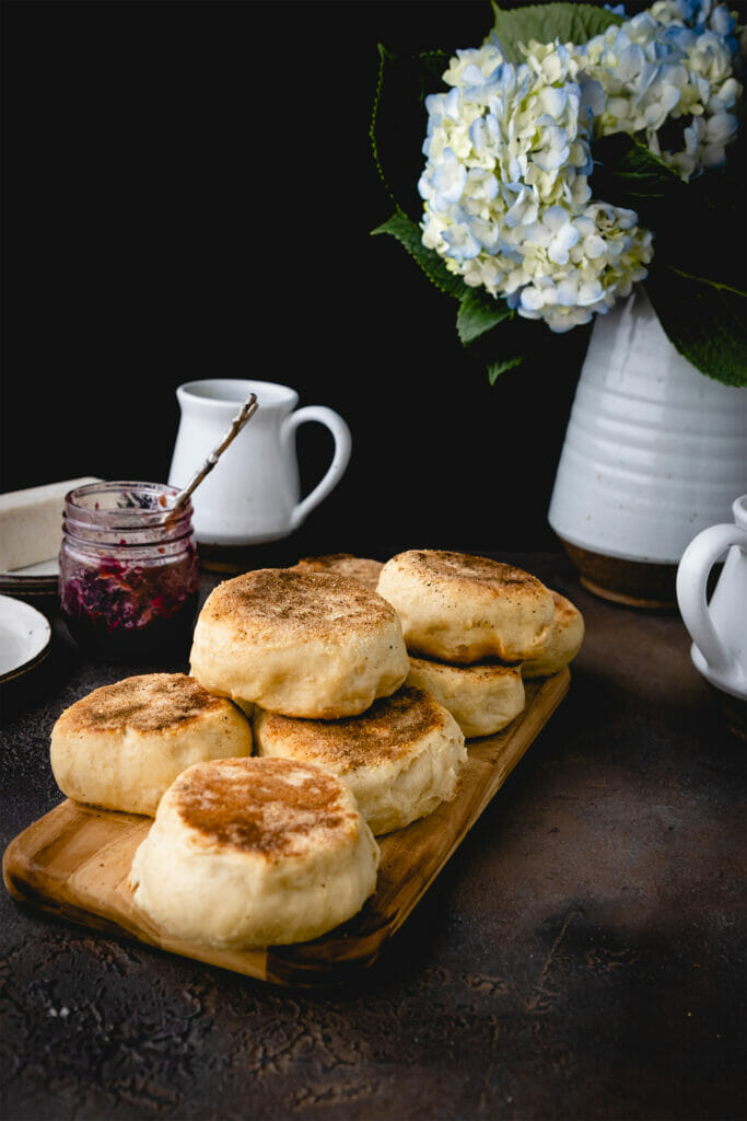 stack of vegan english muffins on a wood board next to a vase of flowers