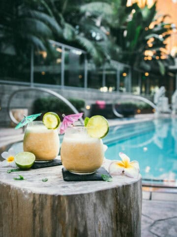two 4 ingredient pina coladas next to a pool on a wooden stand