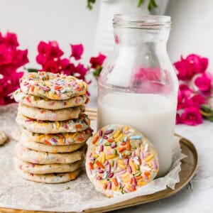 stack of rainbow sprinkle cookies on a tray next to milk