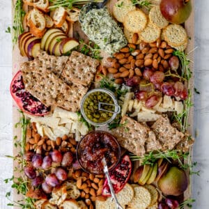 overhead shot of wooden cheese board with a variety of fruits, cheeses, and nuts in a purple green color scheme