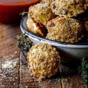 one crispy ravioil bite next to a white dish on a wood board with sauce in the background