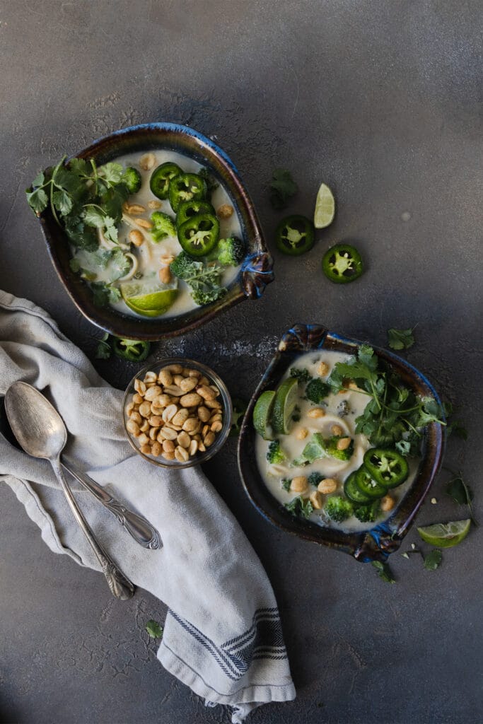 two bowls of spicy peanut noodle soup next to a bowl of peanuts