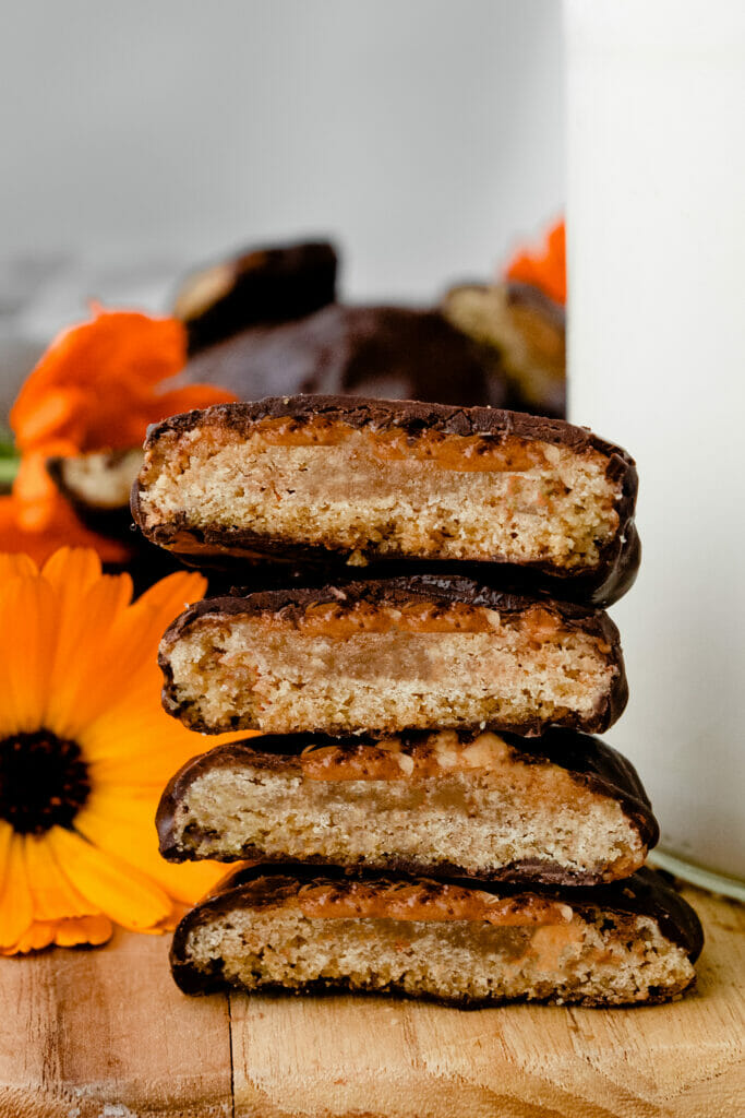 close up of stack of peanut butter patty tagalong cookies on a wooden board, cut open so you can see the layers of cookie and peanut butter. Coated in chocolate