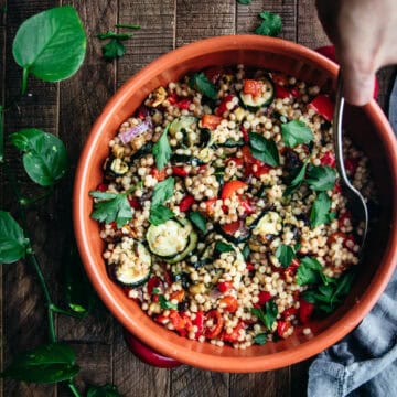 hand with a spoon reaching into a bowl of roasted ratatouille couscous on a wooden table