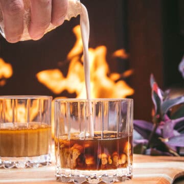 close up of hand pouring coconut cream into a vegan pumpkin spice white russian in front of a roaring fire