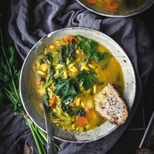 bowl of vegan chickpea orzo soup on a table with a grey cloth and a piece of multigrain bread