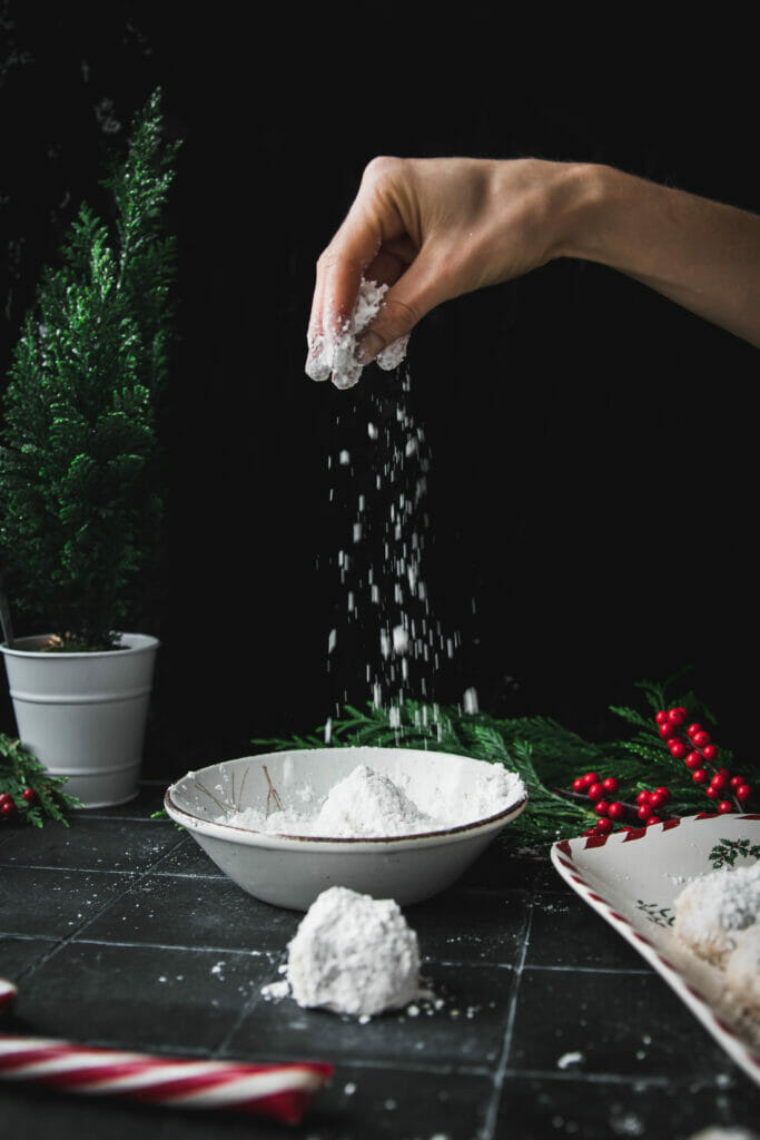 hand pouring powdered sugar onto a toasted walnut snowball cookie