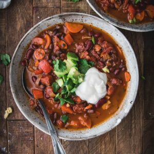 bowl of easy vegan chili topped with green onions, avocado, and sour cream on a wooden table