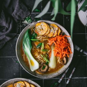 close up of a bowl of vegan sesame miso ramen on a black table next to a vase of leaves