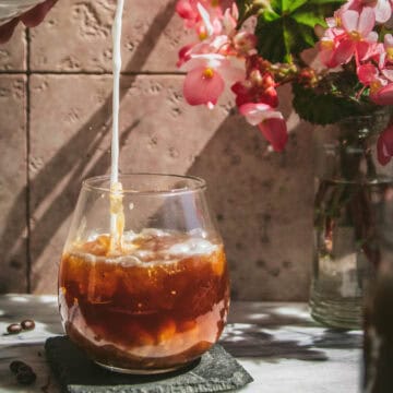 milk pouring into a glass of cinnamon cold brew next to a vase of flowers