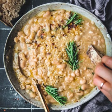 bowl of rosemary garlic braised white beans with a gold spoon and crusty bread